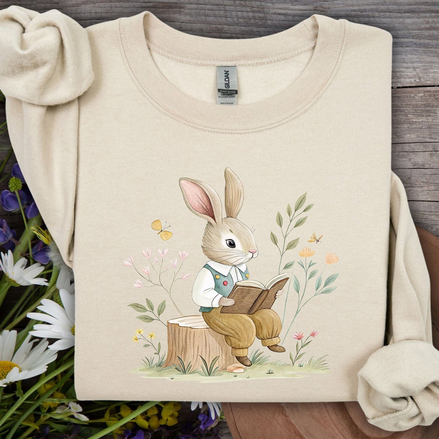 Sand sweatshirt with a watercolor bunny reading on a tree stump, surrounded by spring flowers.