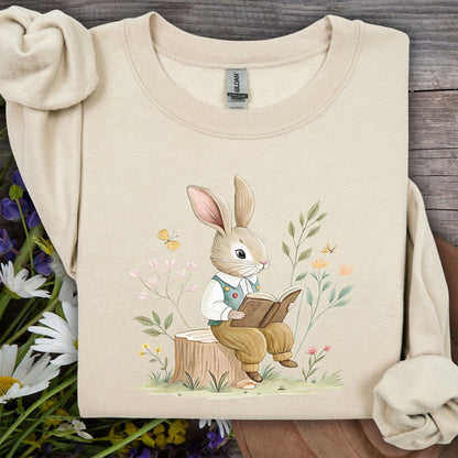 Sand sweatshirt with a watercolor bunny reading on a tree stump, surrounded by spring flowers.