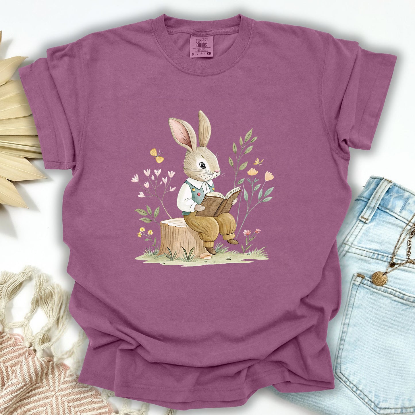 Berry S t-shirt with a watercolor bunny reading on a tree stump amidst flowers.