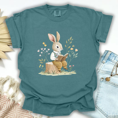 Blue Spruce S t-shirt with a watercolor bunny reading on a tree stump amidst flowers and foliage.