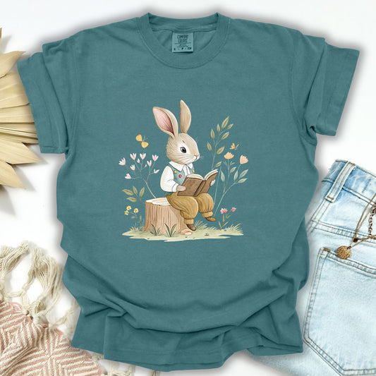 Blue Spruce S t-shirt with a watercolor bunny reading on a tree stump amidst flowers and foliage.