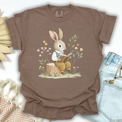 Espresso garment-dyed cotton t-shirt with a watercolor bunny reading on a tree stump, surrounded by flowers.