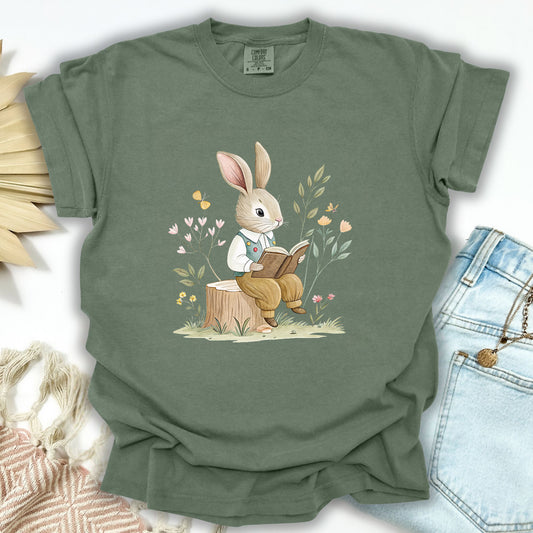 Moss green relaxed-fit t-shirt with a watercolor bunny reading on a tree stump amidst flowers.