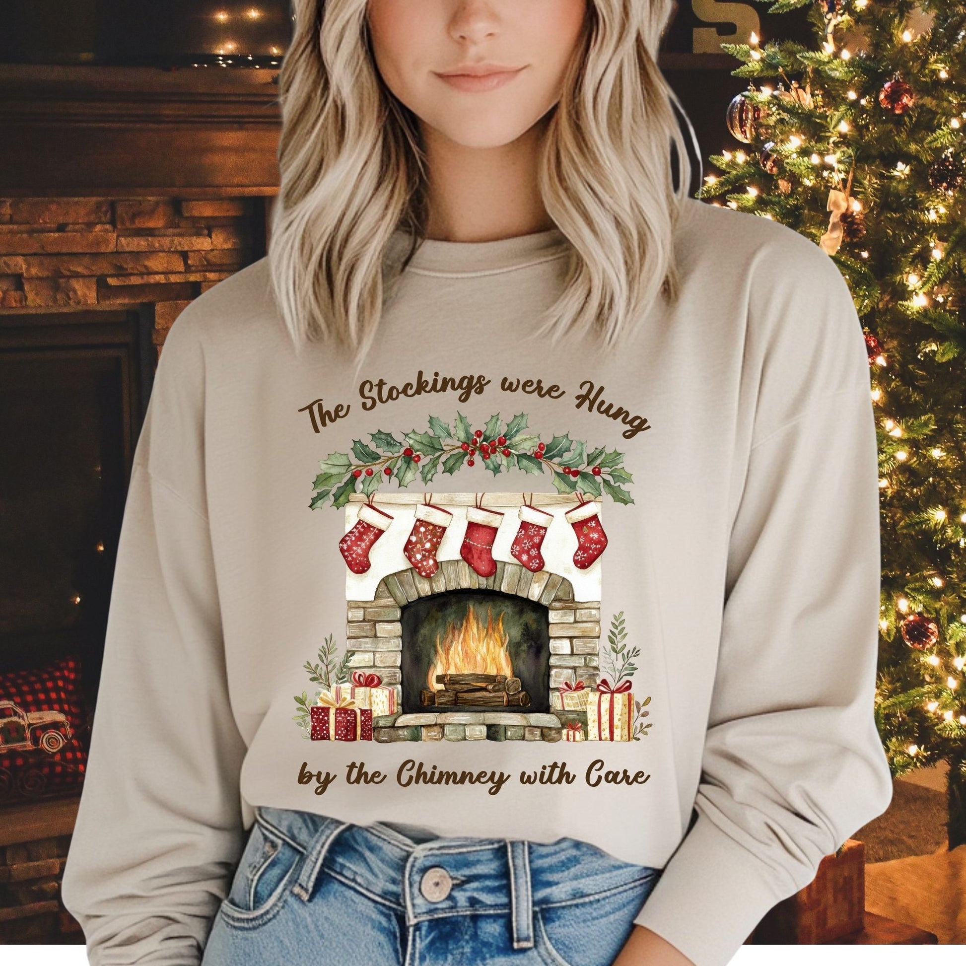 Woman in a beige long sleeve tee with a watercolor fireplace, red stockings, and "The Stockings were Hung" text.