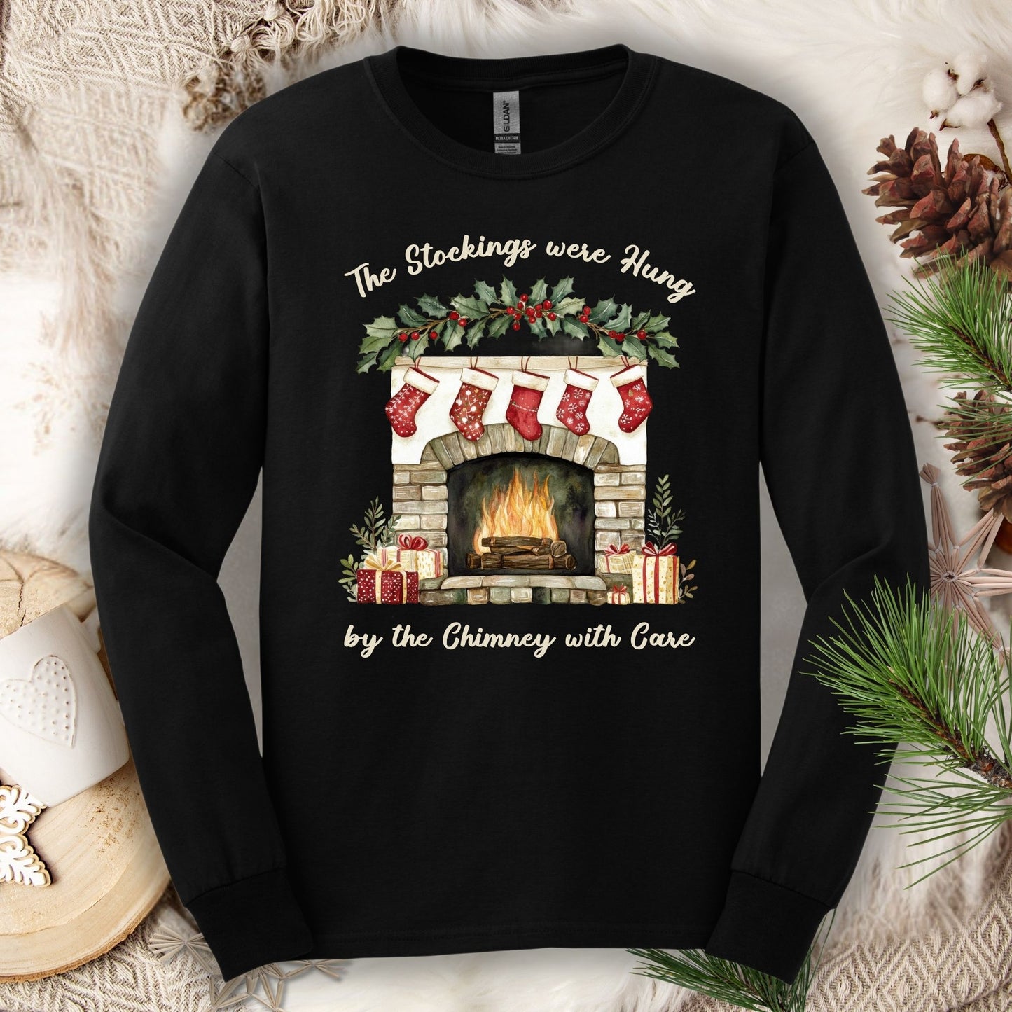 Black long sleeve tee with a watercolor fireplace, red stockings, holly, and "The Stockings were Hung" text.