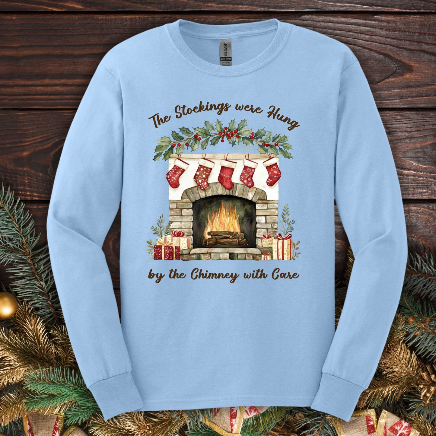 Light blue long sleeve tee with a watercolor fireplace, red stockings, holly, and "The Stockings were Hung" text.