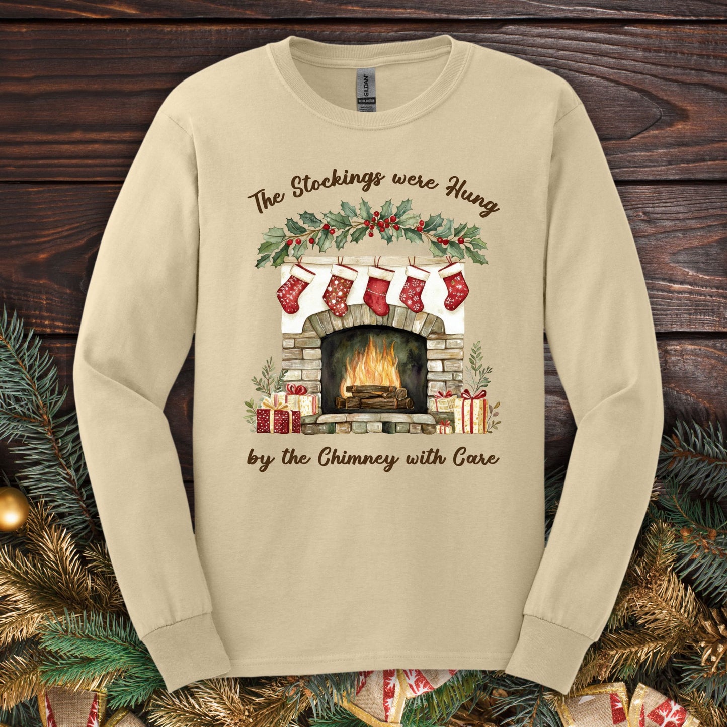 Sand long sleeve tee with a watercolor fireplace, red stockings, holly, and "The Stockings were Hung" text.