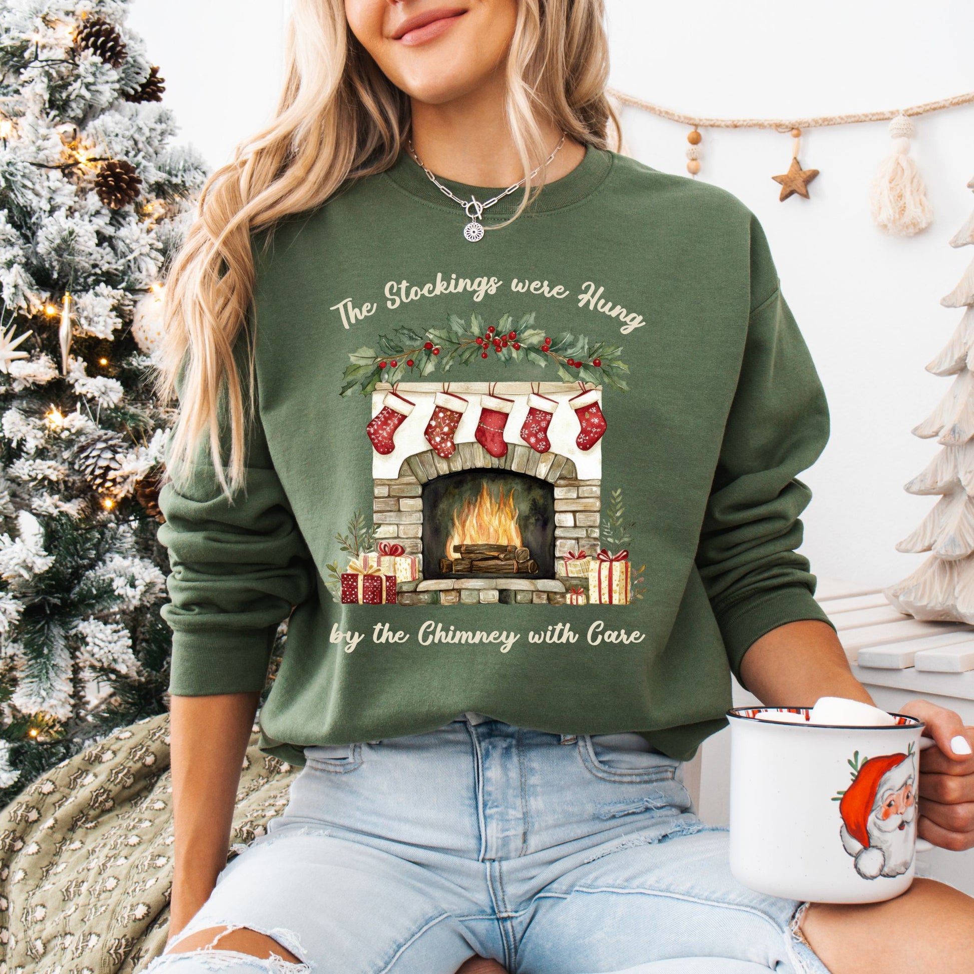 Woman in a green Gildan sweatshirt with a watercolor Christmas fireplace, stockings, and "The Stockings Were Hung" text.