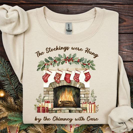 Sand Gildan sweatshirt with watercolor fireplace, red stockings, holly, and "The Stockings were Hung" text.