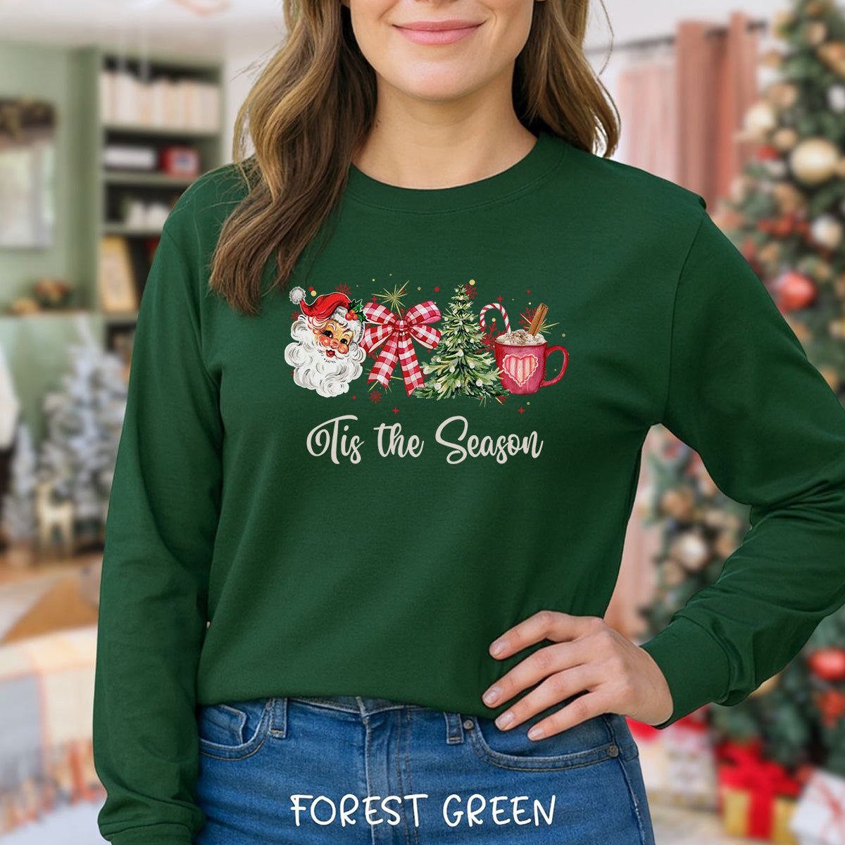 A woman wearing a forest green long-sleeve t-shirt with a Christmas graphic that reads 'Tis the Season'. The graphic features a Santa Claus, a red and white checkered bow, a green Christmas tree, a red mug, and a candy cane.