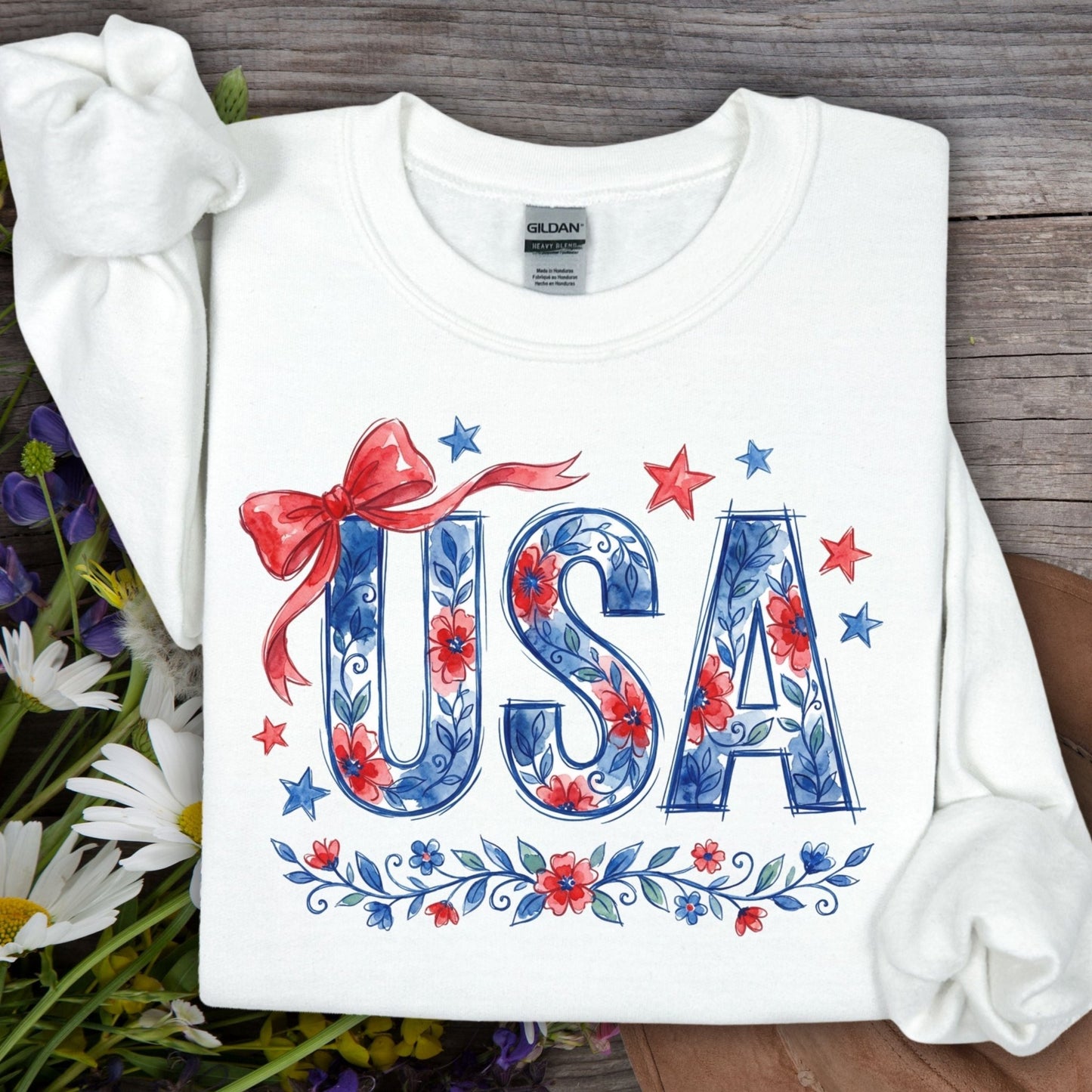 White USA Patriotic Floral Sweatshirt with blue floral lettering, red bow, and stars on a wooden background.