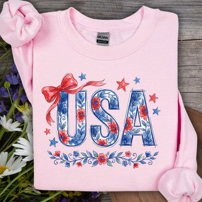 Light pink USA Patriotic Floral Sweatshirt with blue floral lettering, red bow, and stars on wood.