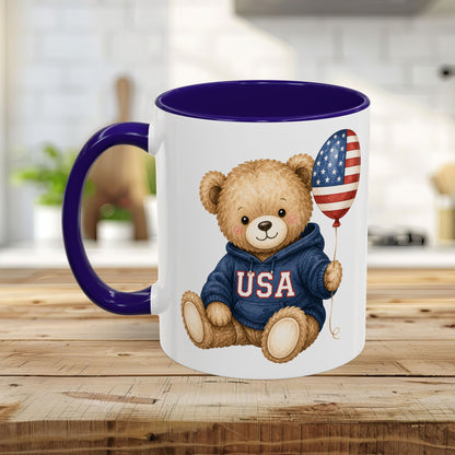White ceramic mug with a purple handle and interior, featuring a teddy bear in a USA hoodie holding a flag balloon.