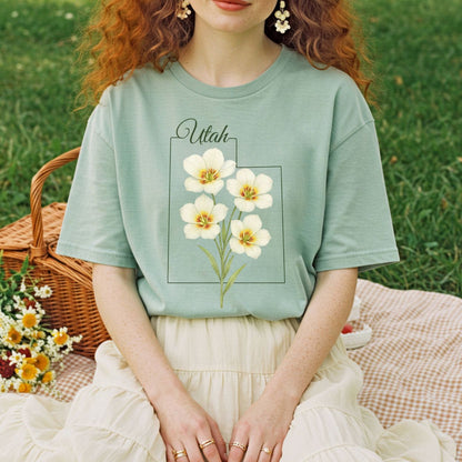Sage green Utah State Flower T-Shirt with watercolor Sego Lilies, worn by a person on a picnic blanket.
