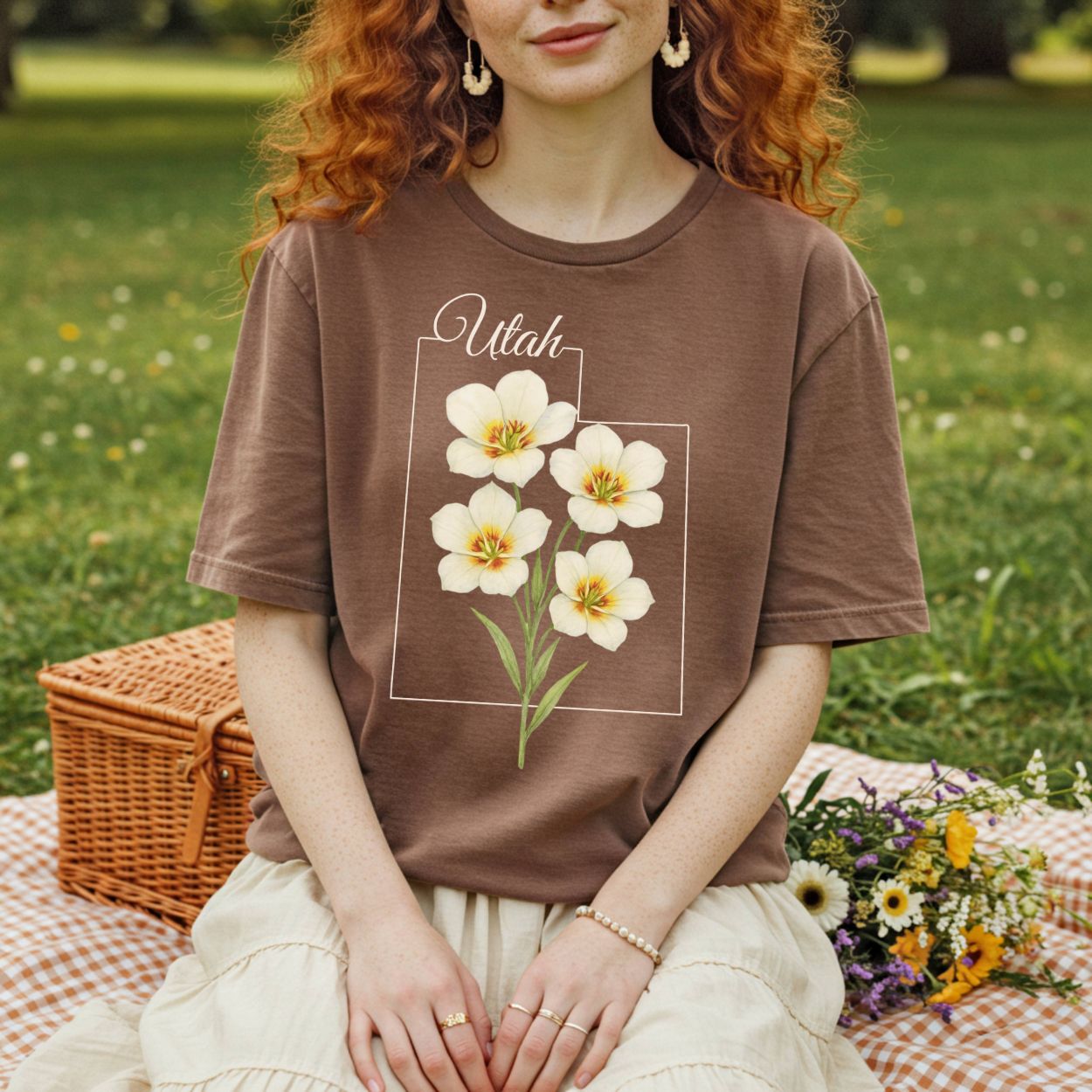 Brown t-shirt with watercolor sego lilies and "Utah" text, worn by a person sitting on a picnic blanket.