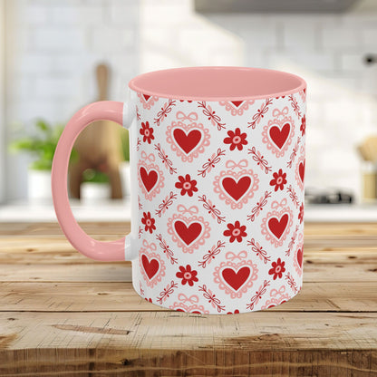 Ceramic mug with a pink handle and interior, featuring a repeating pattern of red hearts, floral motifs, and bows.