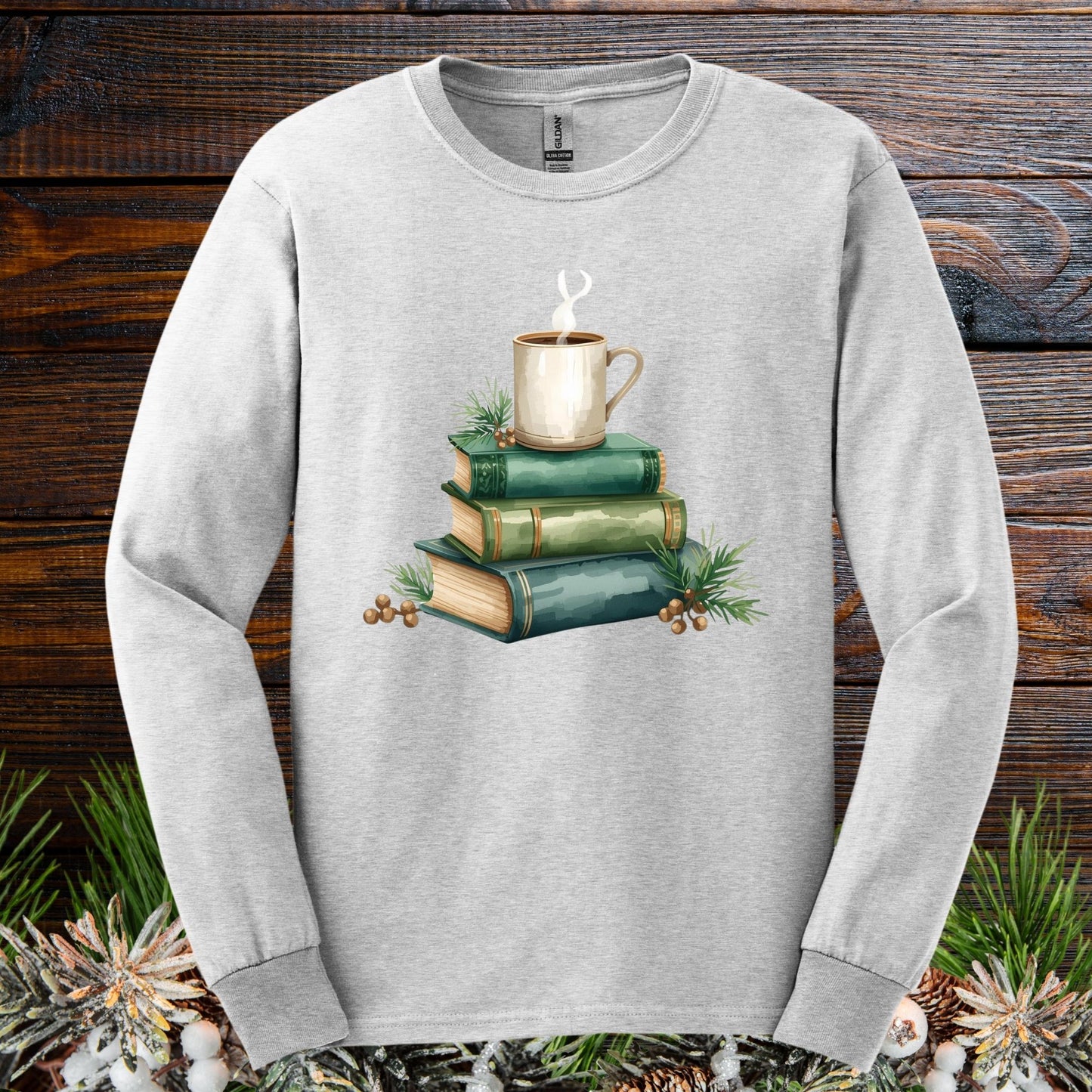 Ash S long sleeve tee with a watercolor design of vintage green books, a steaming coffee mug, and pine accents.