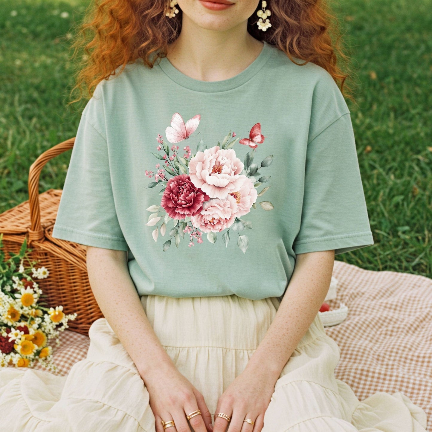 Sage green relaxed-fit t-shirt with a watercolor peony and butterfly bouquet design, worn by a person sitting on a picnic blanket.