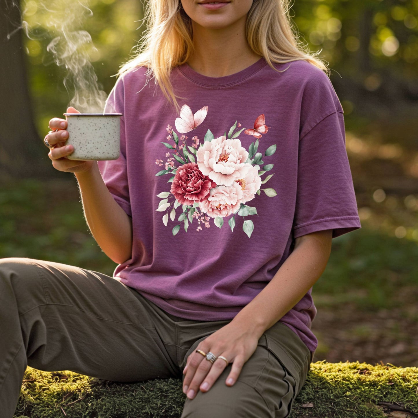 Woman in a relaxed-fit, plum garment-dyed tee with a watercolor peony and butterfly bouquet, holding a steaming mug.