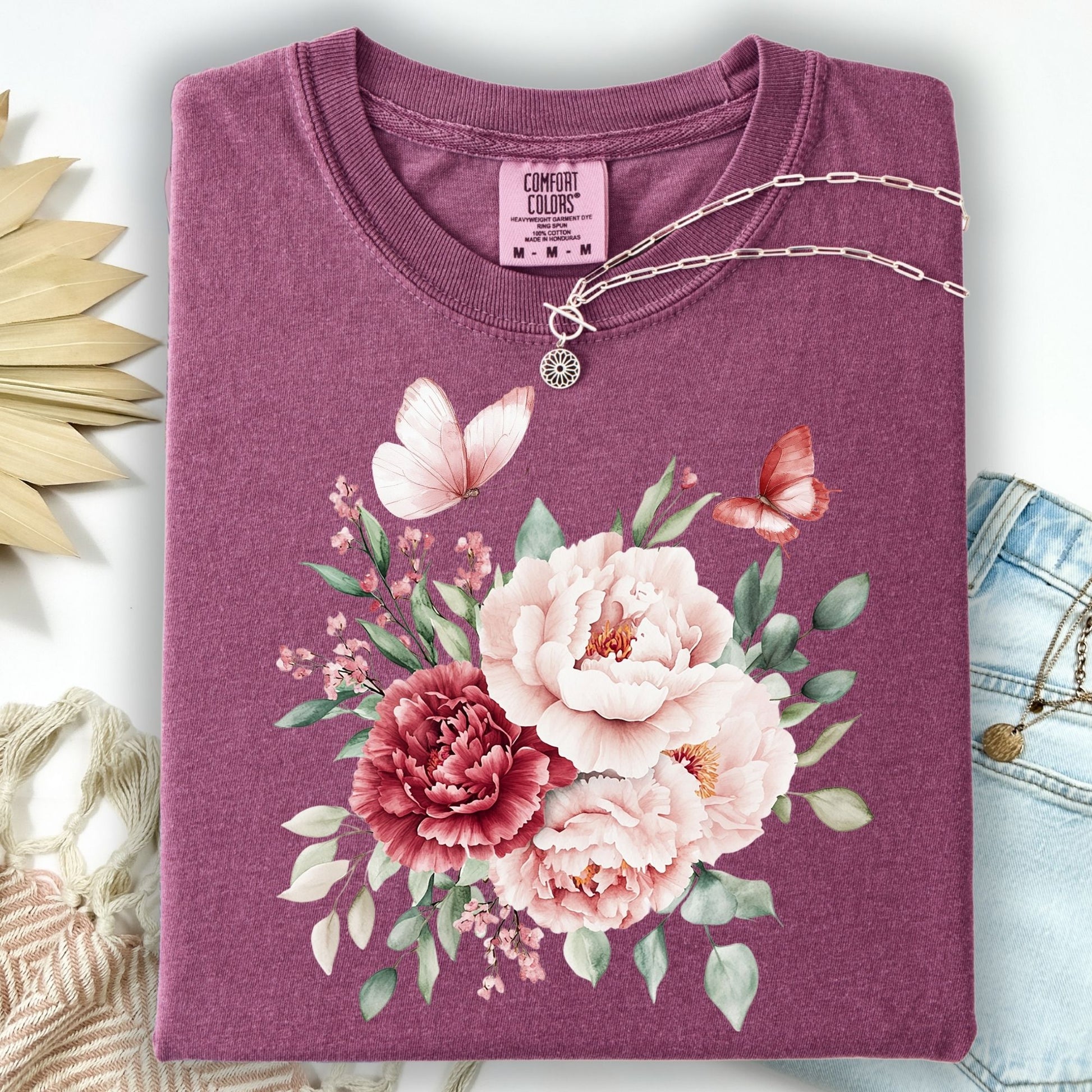 Berry garment-dyed cotton t-shirt with a vintage peony and butterfly watercolor design, folded with a silver necklace.