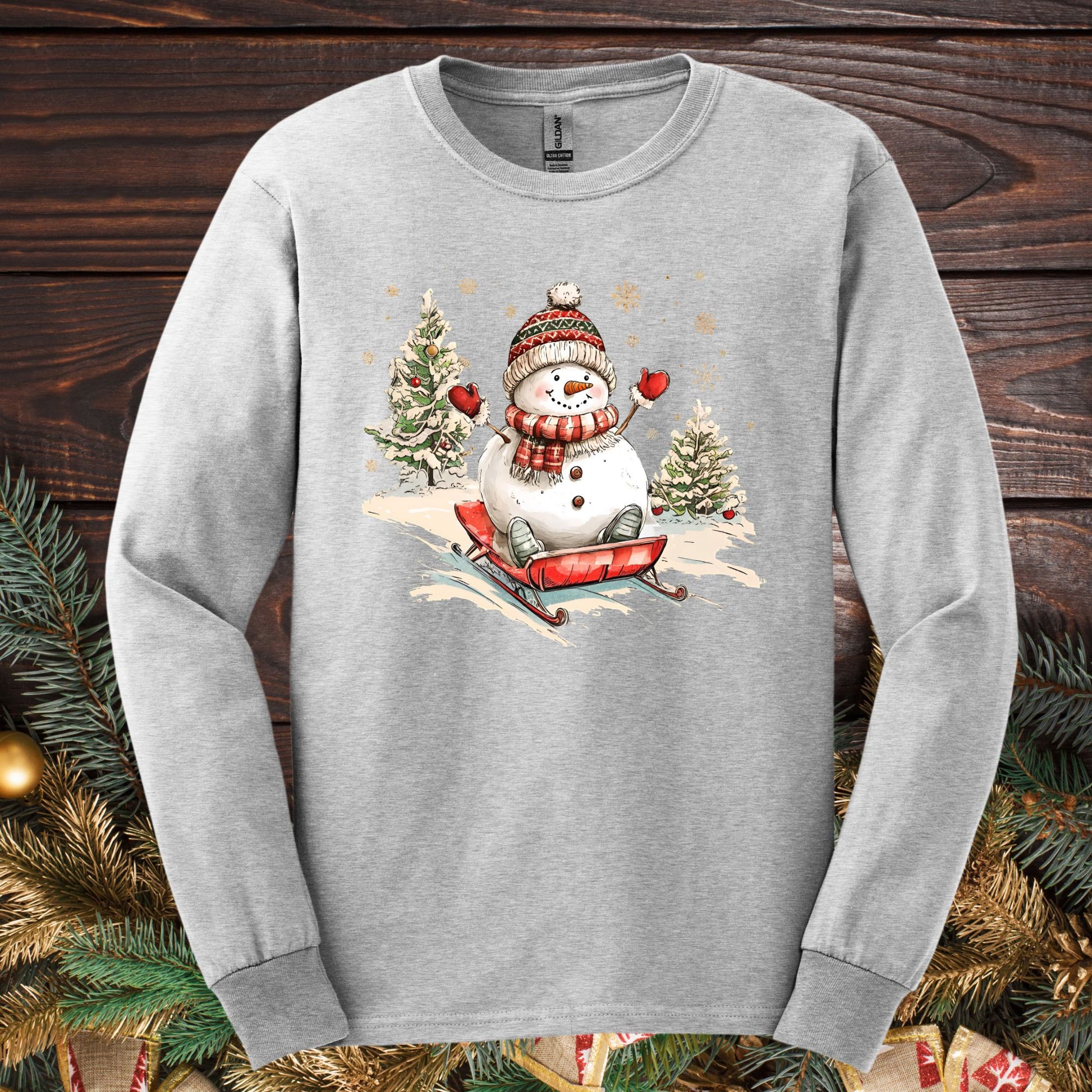Ash Gildan long sleeve tee with a vintage snowman sledding design, surrounded by pine boughs.