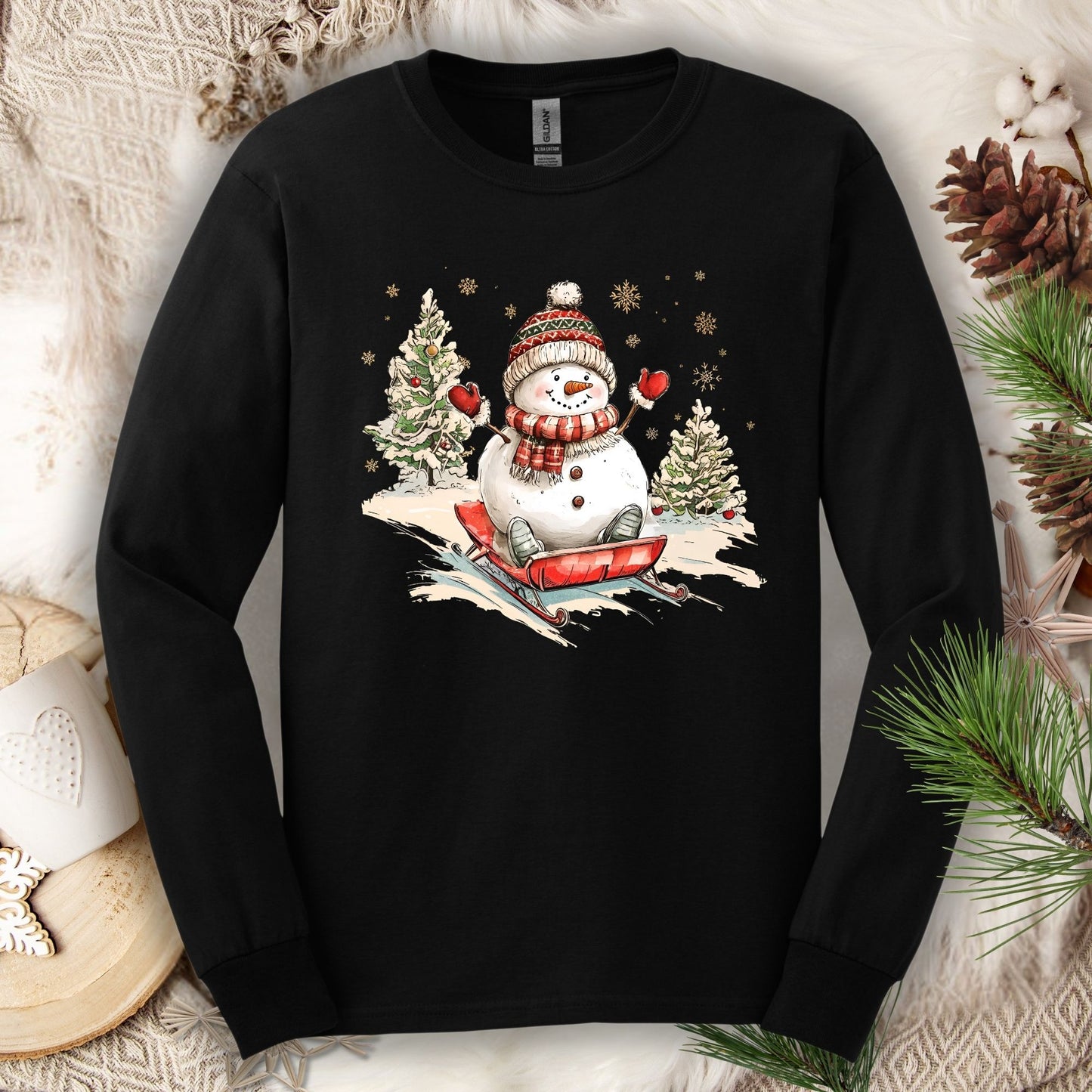 Black Gildan long sleeve tee with a vintage snowman sledding design, surrounded by snowy trees and snowflakes.