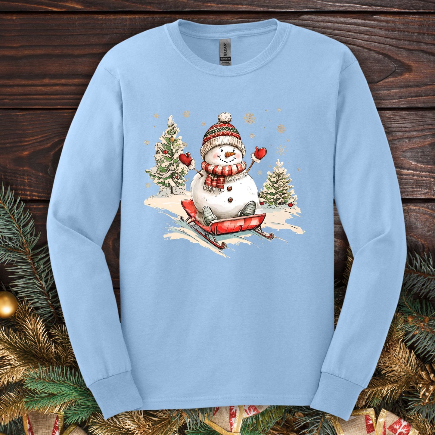 Light blue Gildan long sleeve tee with a vintage snowman sledding design, surrounded by snowy trees and snowflakes.