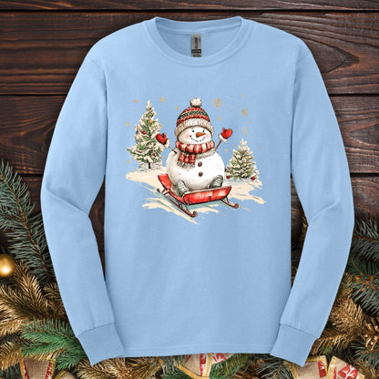 Light blue Gildan long sleeve tee with a vintage snowman sledding design, surrounded by snowy trees and snowflakes.