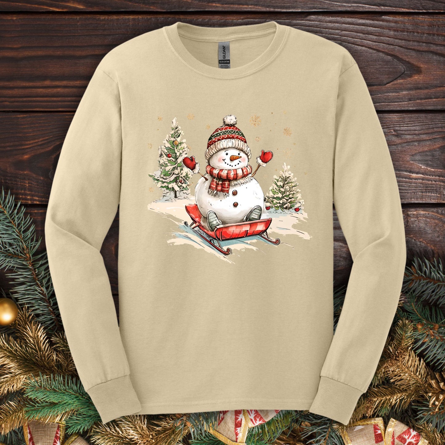 Sand long sleeve tee with a vintage snowman sledding design, rosy cheeks, scarf, and winter trees.