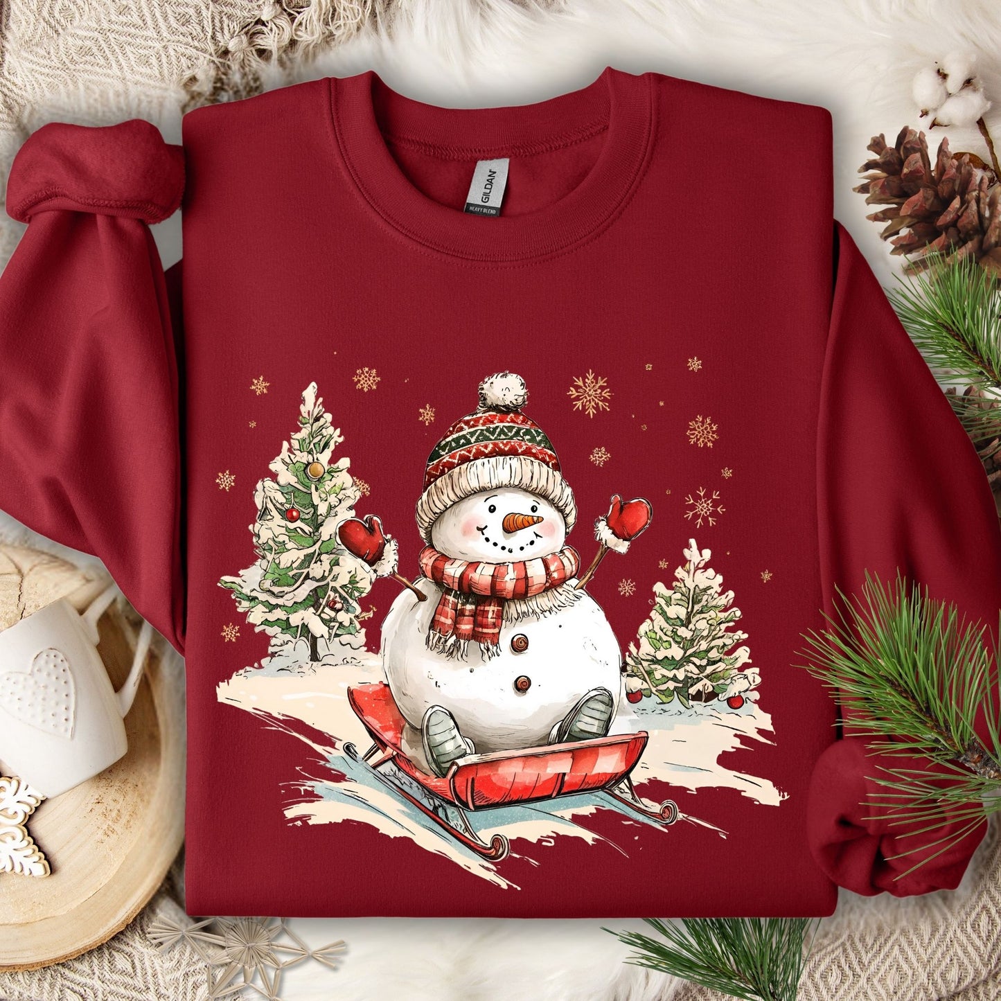 Cardinal Red Gildan sweatshirt with a vintage snowman sledding design, surrounded by winter trees and snowflakes.