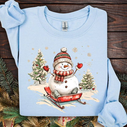Light blue Gildan sweatshirt with a vintage snowman sledding design, surrounded by snowy trees and snowflakes.