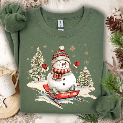 Military Green Gildan sweatshirt with a vintage snowman sledding design, surrounded by snowy trees and snowflakes.