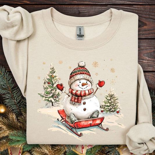 Sand Gildan sweatshirt with a vintage snowman sledding design, surrounded by trees and snowflakes.