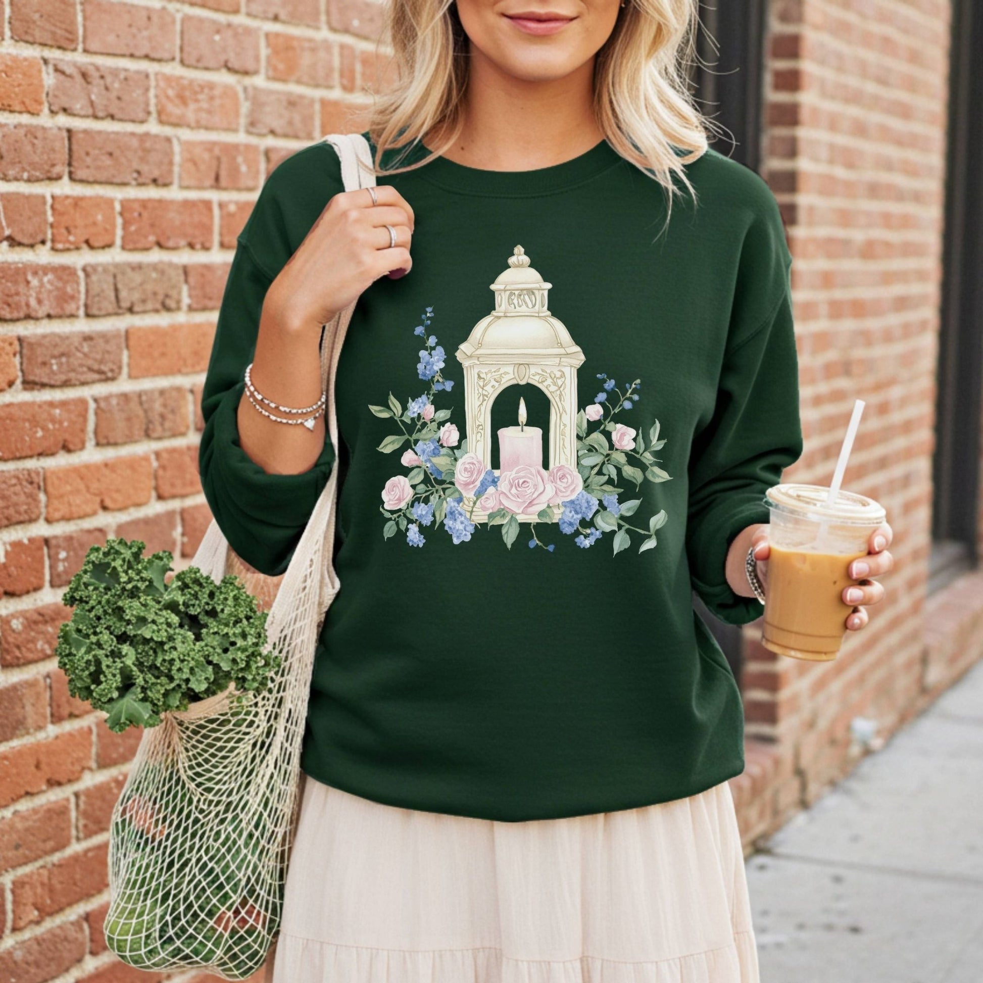 Woman in a dark green crewneck sweatshirt with a vintage lantern and watercolor roses, holding coffee and a market bag.