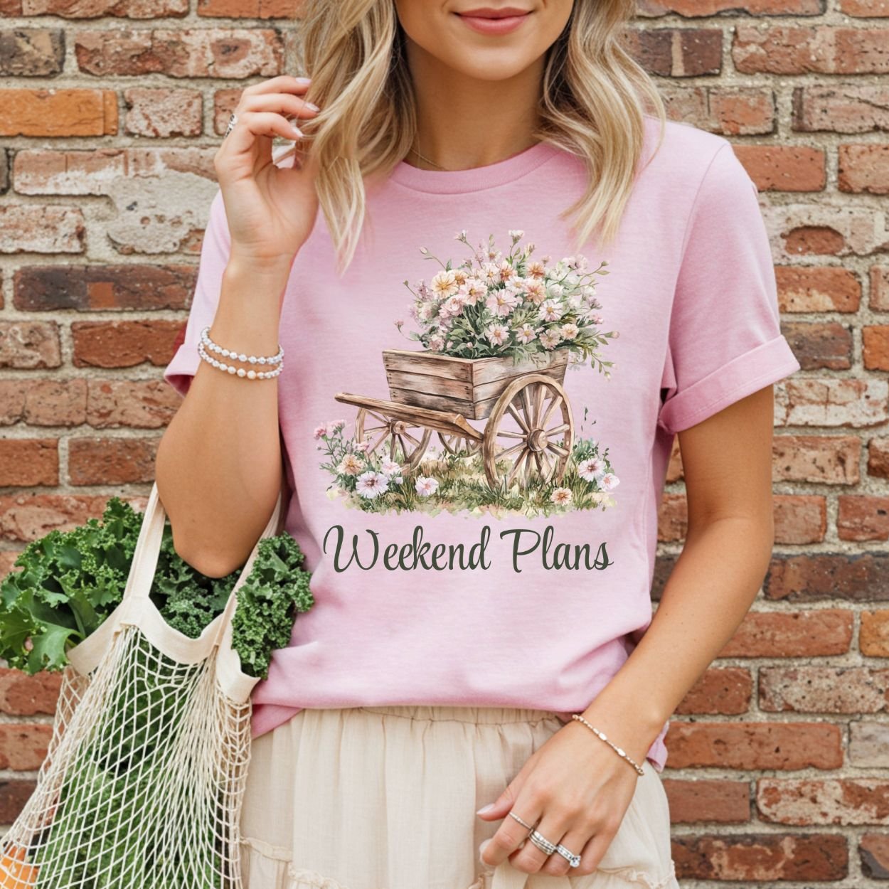 Pink garment-dyed t-shirt with a watercolor gardening wagon overflowing with carnations and "Weekend Plans" text.