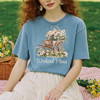 young woman wearing a blue t-shirt with a floral design and 'Weekend Plans' text, sitting outdoors.