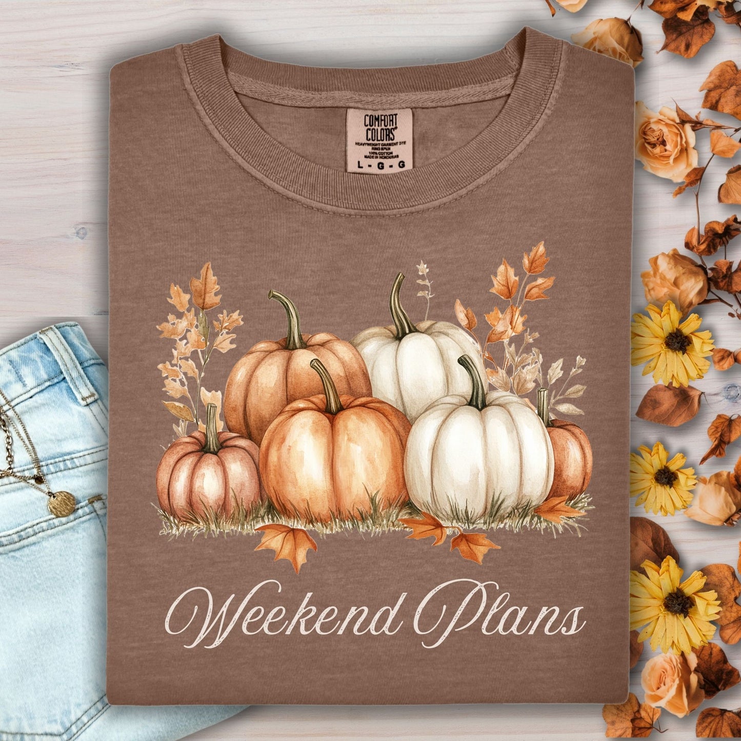 Espresso heavyweight cotton t-shirt with watercolor pumpkins and "Weekend Plans" text, folded beside jeans and fall decor.
