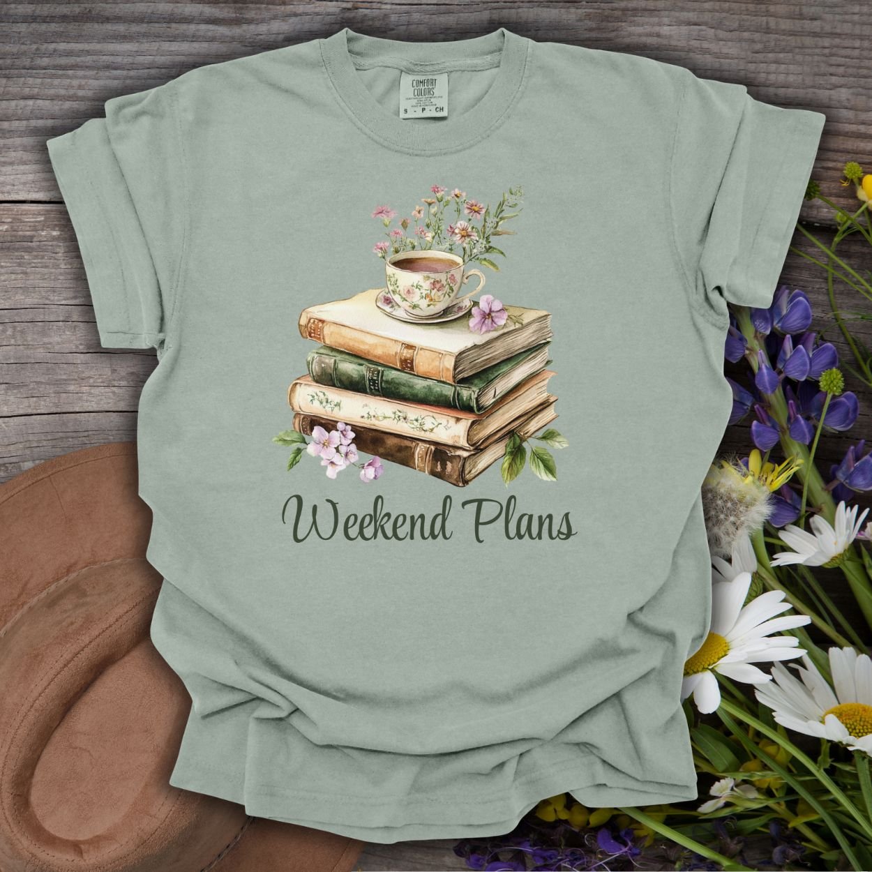 Bay green garment-dyed cotton t-shirt with a watercolor stack of books, teacup, flowers, and "Weekend Plans" text.