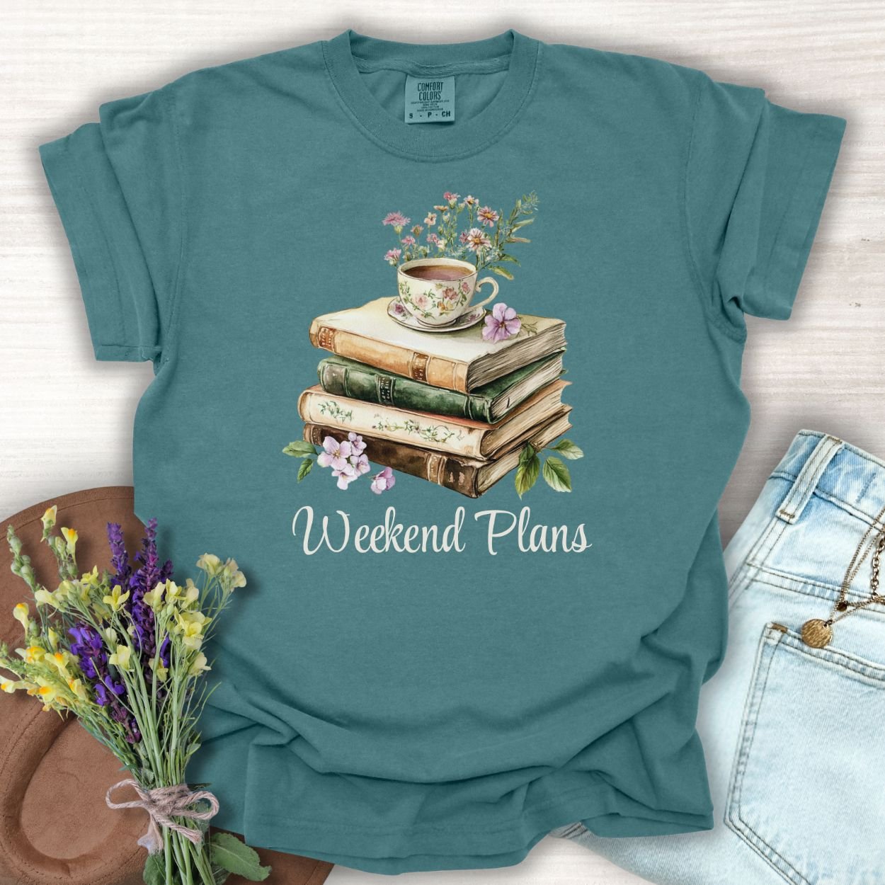 Blue Spruce garment-dyed cotton t-shirt with watercolor books, teacup, flowers, and "Weekend Plans" text.