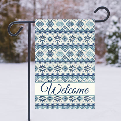 Blue Fair Isle pattern garden flag with "Welcome" script, hanging on a black stand in a snowy setting.
