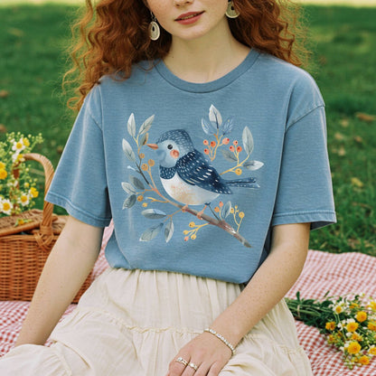 Garment-dyed blue t-shirt with a whimsical watercolor bluebird on a branch with berries, worn by a woman.