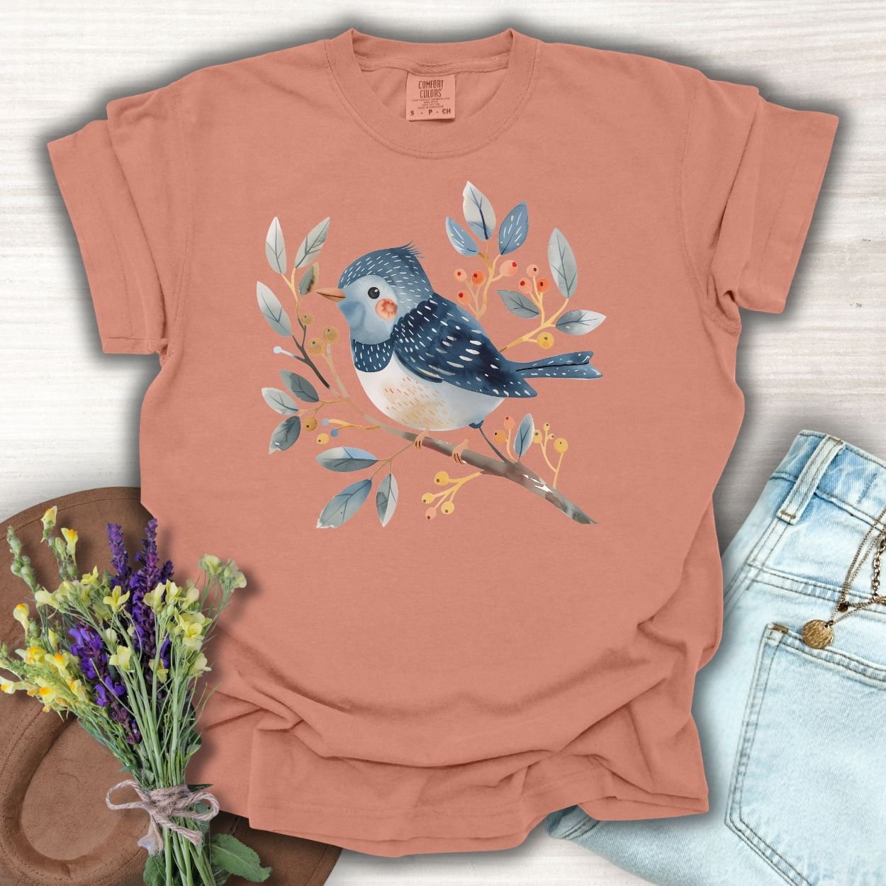 Terracotta S t-shirt with a whimsical bluebird on a branch, surrounded by blue leaves and red/orange berries.