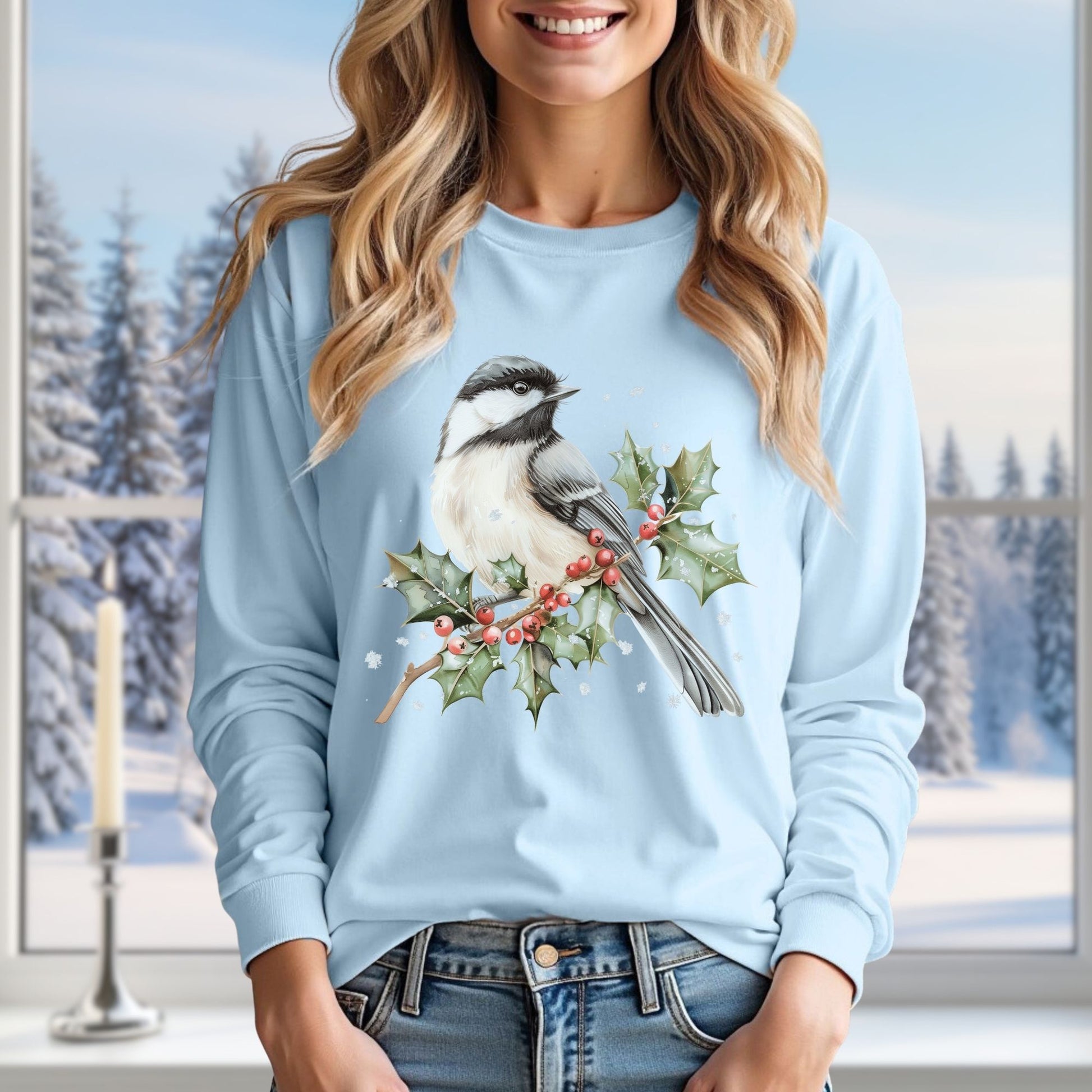 Light blue long sleeve tee with a watercolor chickadee on a snowy holly branch, worn by a smiling woman.