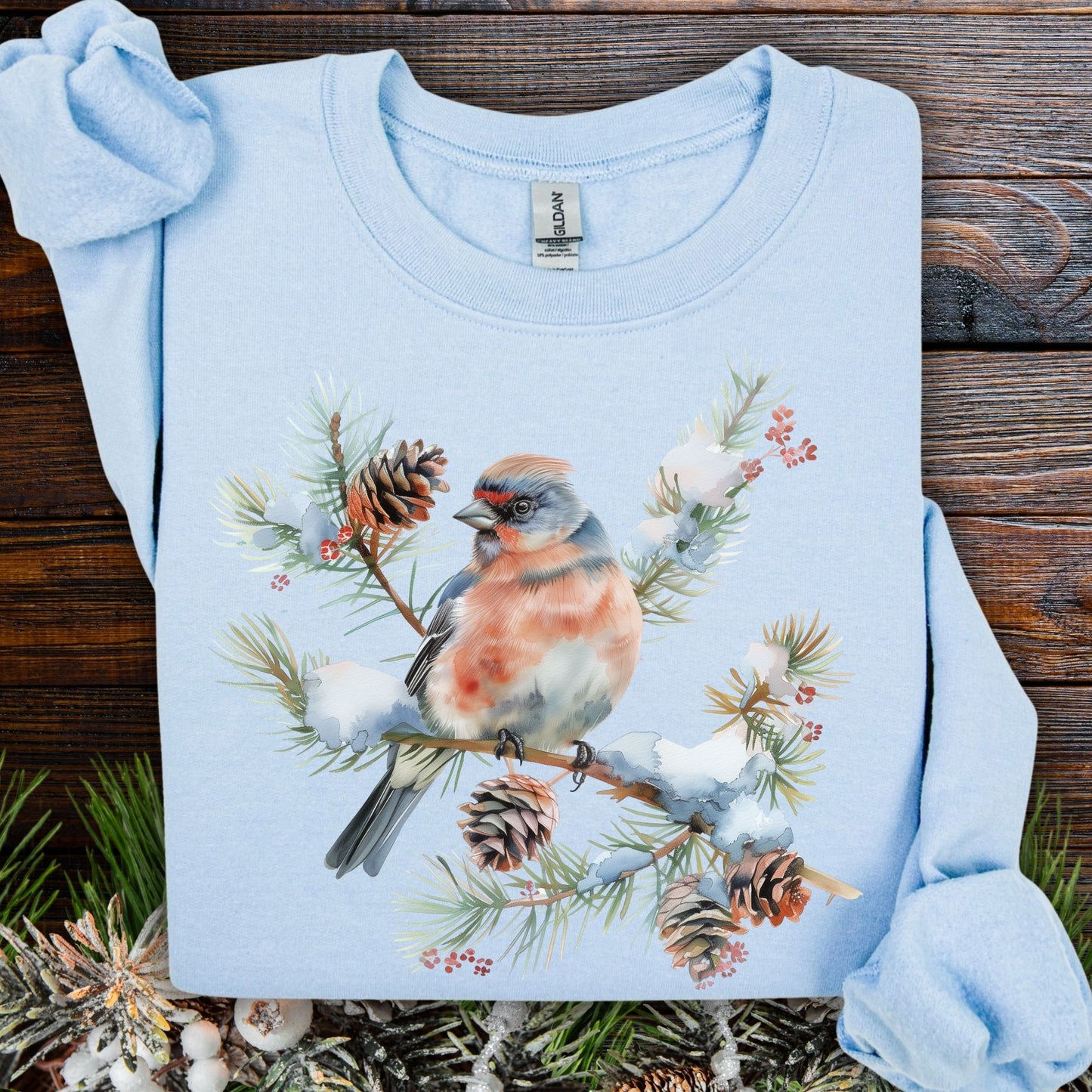 Light blue sweatshirt with a crested songbird on a snowy evergreen branch with pinecones and berries.