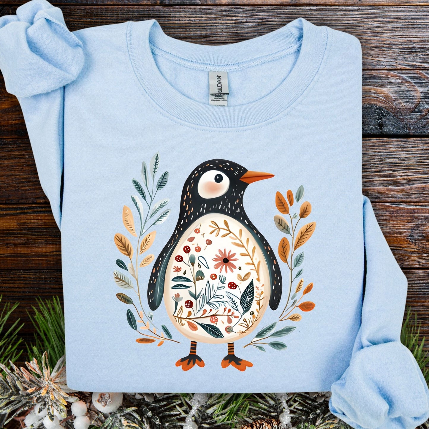 Light blue sweatshirt with folk art penguin, botanical patterns, orange beak, and striped legs.