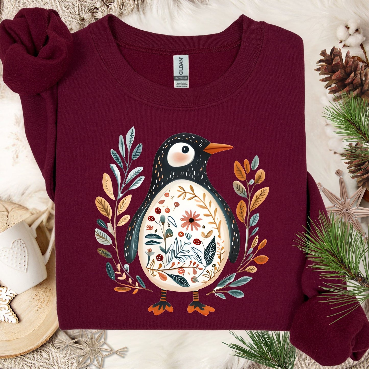 Maroon sweatshirt with a folk art penguin adorned with botanical patterns, flowers, and an orange beak.