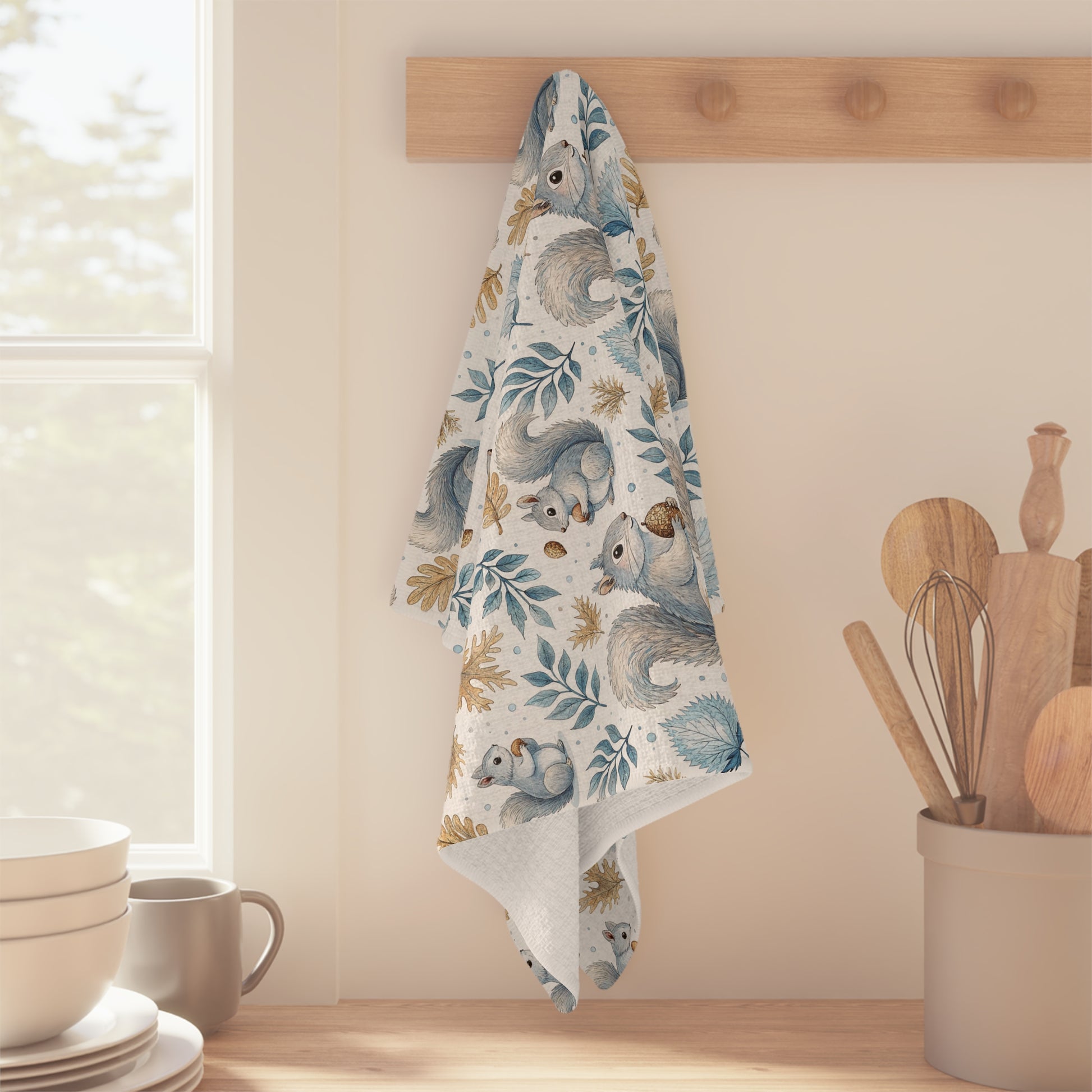 Microfiber tea towel with watercolor squirrels, blue leaves, and ochre acorns, hanging on a wooden rack.