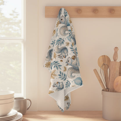 Microfiber tea towel with watercolor squirrels, blue leaves, and ochre acorns, hanging on a wooden rack.