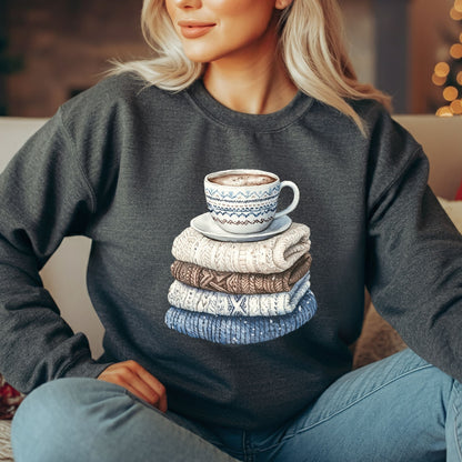 Blonde woman wearing a dark gray crewneck sweatshirt with a watercolor design of stacked knit sweaters and a coffee mug.