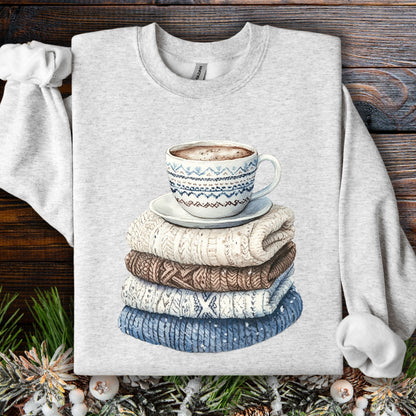 Watercolor-style graphic of a patterned mug on a stack of folded knit sweaters on a light gray sweatshirt.